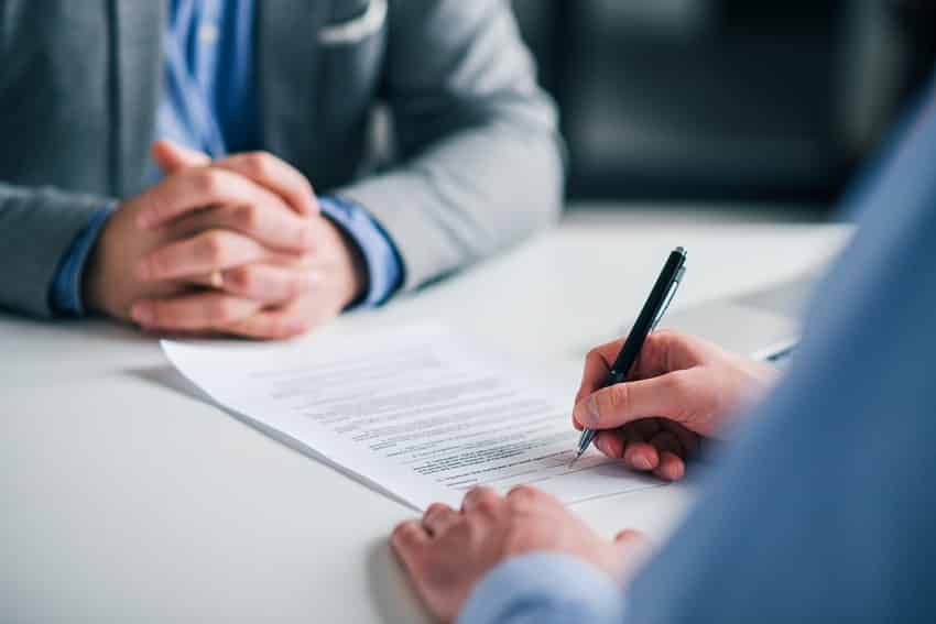 Arbeitsvertrag unterzeichnen Person unterschreibt einen Vertrag, während eine zweite Person mit gefalteten Händen zusieht