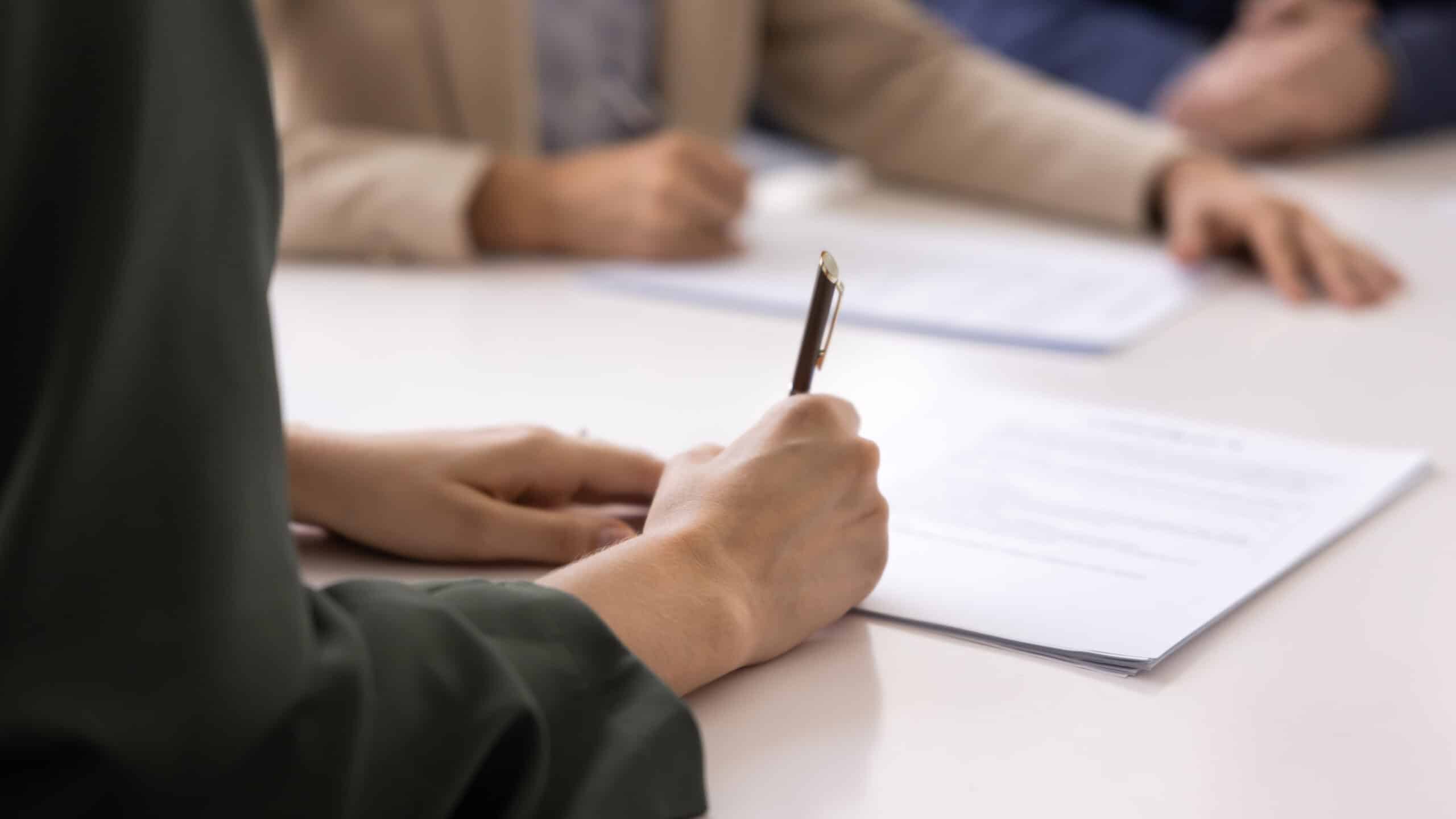 Person mit Stift über einem Vertragsdokument im Beratungsgespräch am Tisch Person mit Stift über einem Vertragsdokument im Beratungsgespräch am Tisch