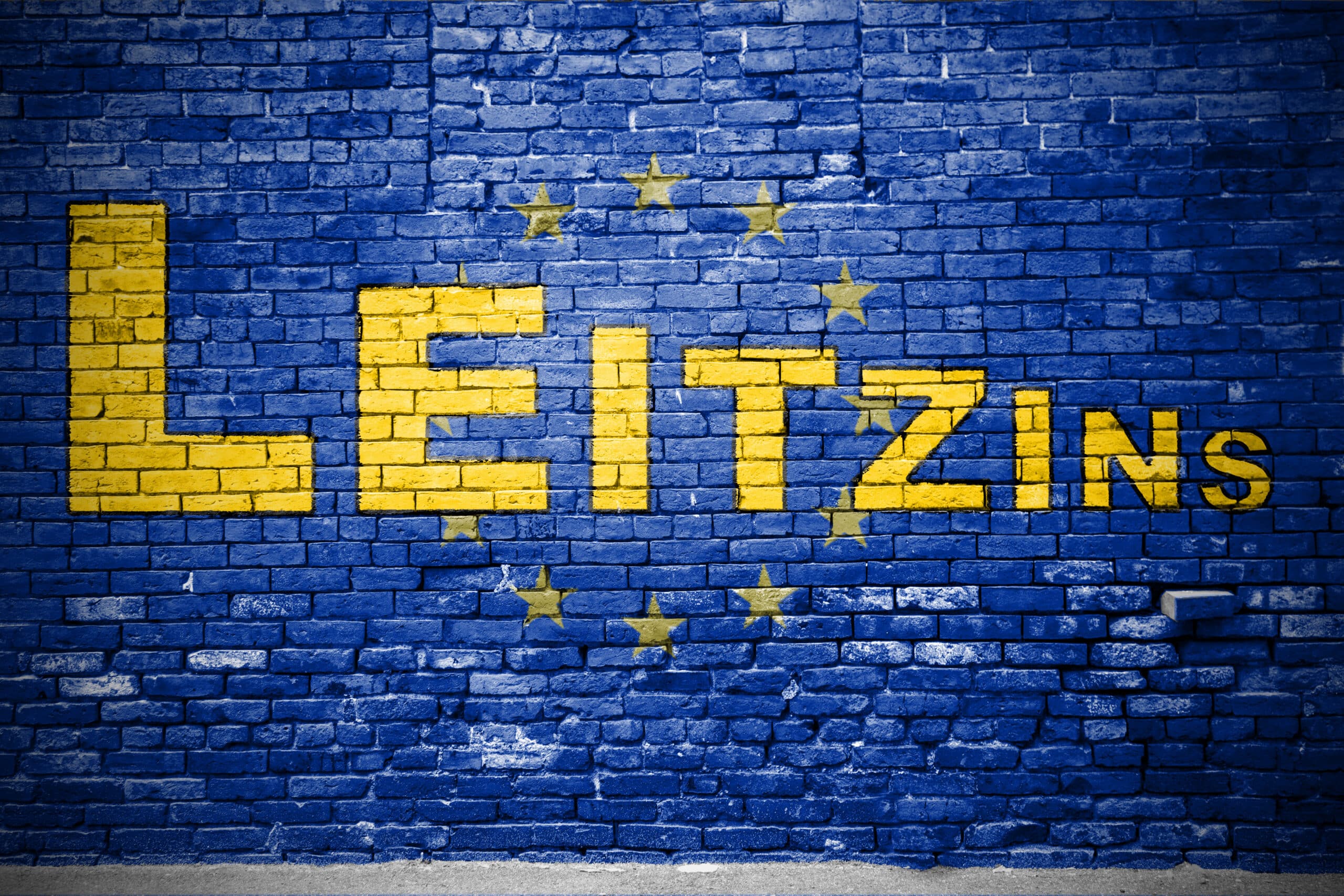 Leitzins der EZB: Symbolbild EU-Flagge Schriftzug Leitzins in gelben Buchstaben auf einer Backsteinwand im Design der EU-Flagge