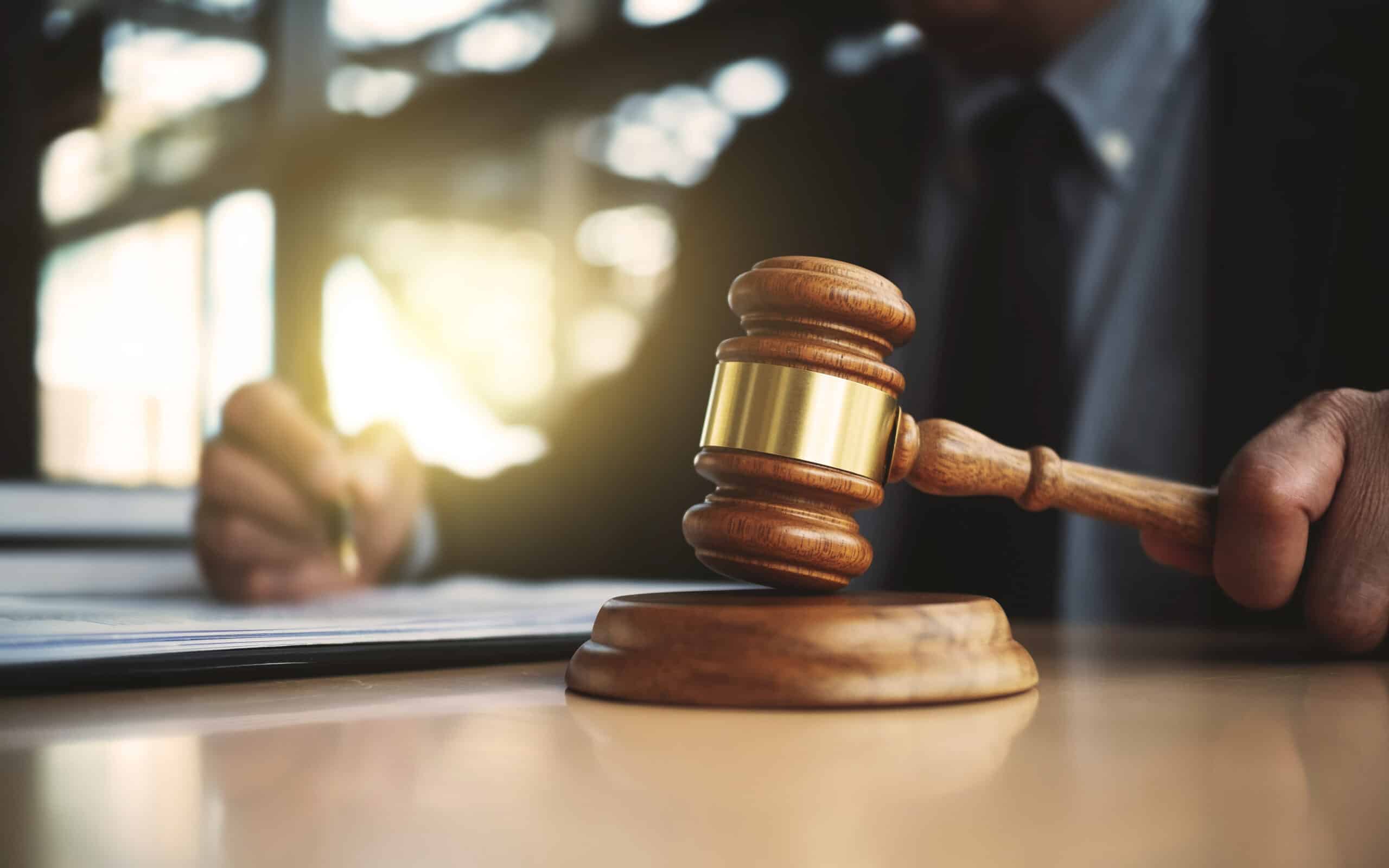 Zwangsversteigerung: Zuschlag durch das Gericht Richterhammer mit goldenem Ring auf einem Holzsockel, im Hintergrund eine Person am Tisch